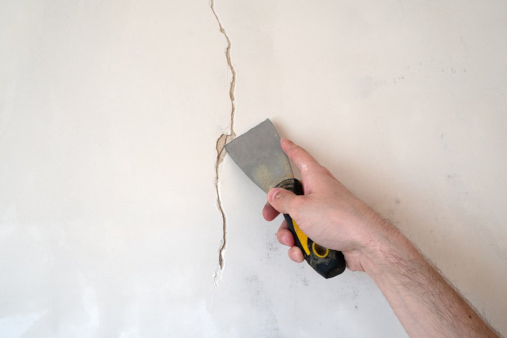 Een close-up van een scheur in stucwerk op een muur, met gereedschap en reparatiemateriaal op de achtergrond.