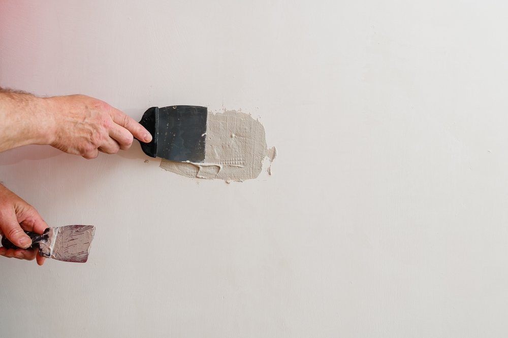 Een close-up van een hand die een groot gat in de muur vult met reparatiemateriaal, met gereedschap en reparatiemateriaal op de achtergrond.