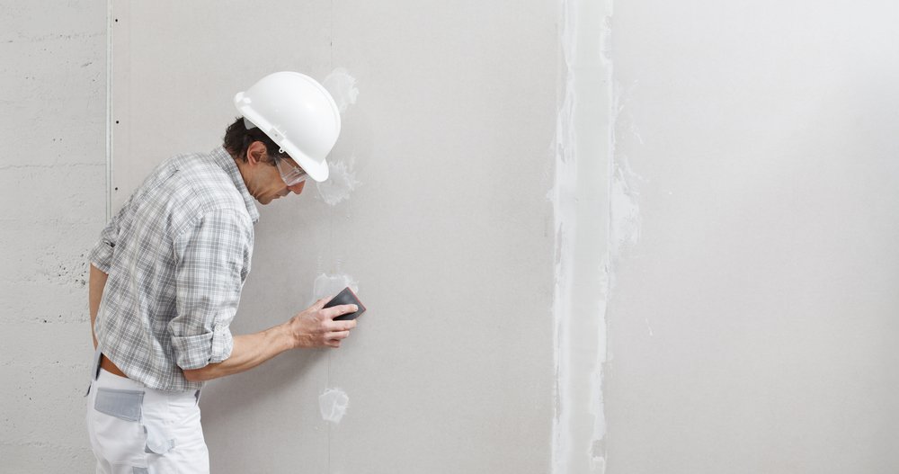 Een close-up van een hand die stucwerk repareert op een muur met een plamuurmes en vulmiddel, met gereedschap en reparatiemateriaal op de achtergrond.