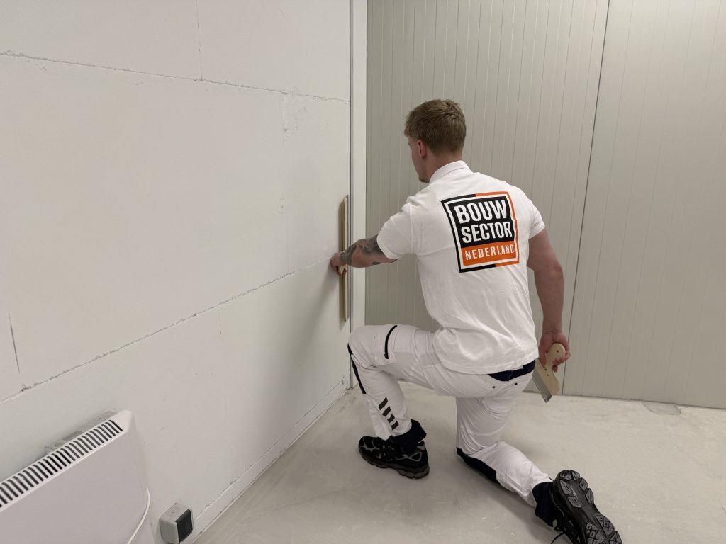 Stukadoor van Bouwsector Nederland zit geknield bij een wand naast een verwarmingspaneel en zet een spaan tegen de wand